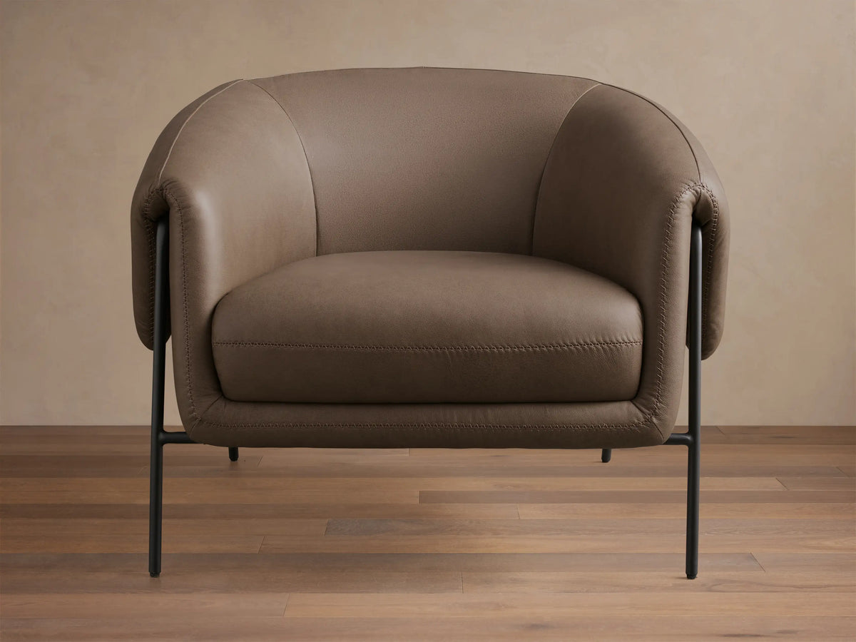 Front view of the taupe Linus Leather Chair, with curved leather upholstery, visible stitching, and black metal legs.