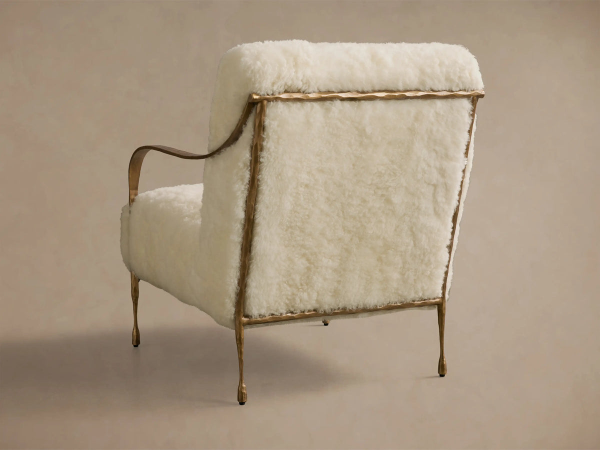 Rear view of the cream Gwen Shearling Chair with a hammered brass frame, curved armrest, and tapered legs.