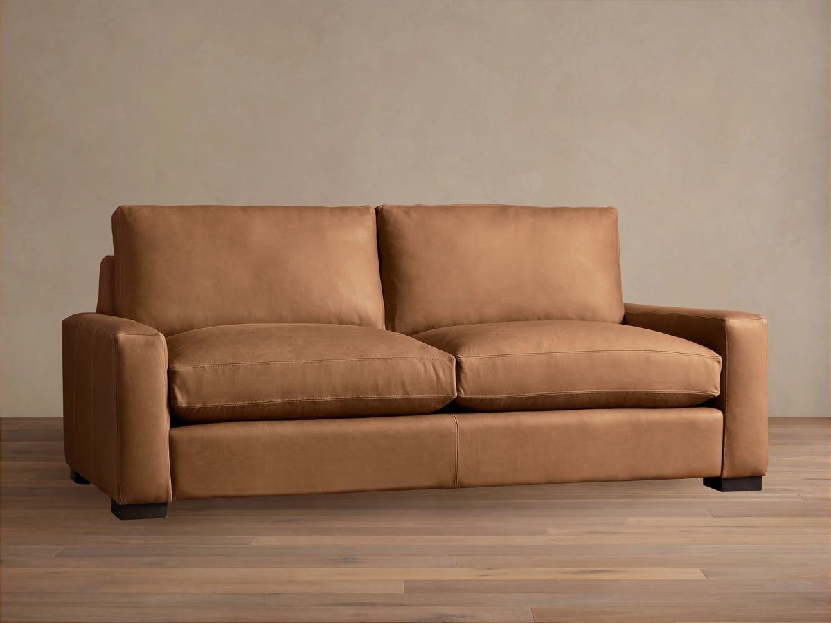 Tan Remington Leather Sofa with plush backrests and seat cushions, straight arms, and dark wooden legs, on a wooden floor.