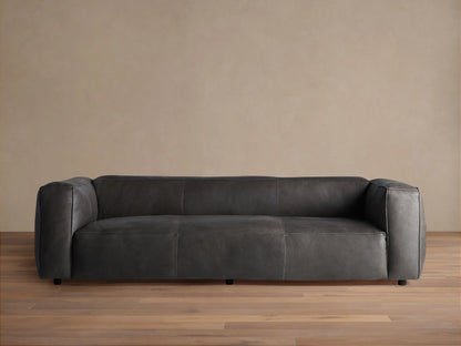 Madrone Leather Sofa, dark grey with modern, blocky arms & visible stitching. It floats on recessed legs against a neutral wall.