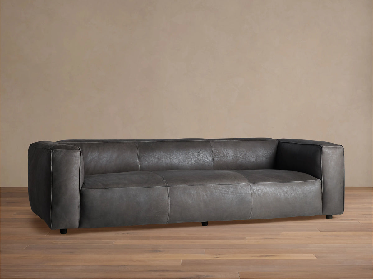 Madrone Leather Sofa, dark grey with modern, blocky arms & visible stitching. It floats on recessed legs against a neutral wall.