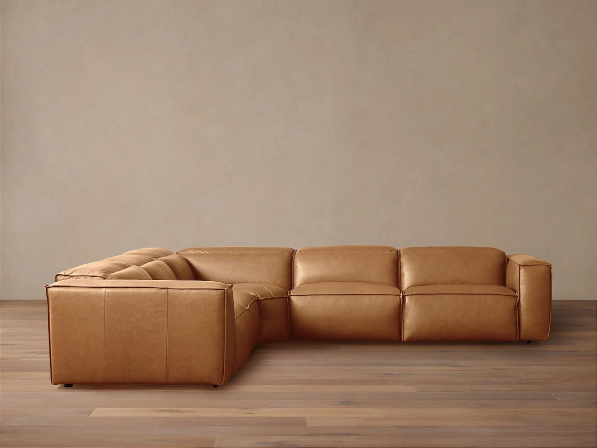 Tan leather Coburn Five-Piece Motion Corner Sectional. L-shaped sofa with plush cushions on wooden floor, beige wall.