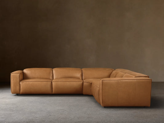 Tan leather Coburn Five-Piece Motion Corner Sectional. L-shaped sofa with plush cushions on wooden floor, beige wall.