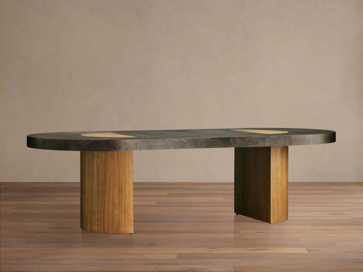 Oval Acacius Dining Table with a dark, naturally oxidized steel top and two cylindrical wooden legs on a wooden floor.