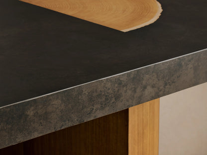 Oval-shaped Acacius Dining Table with a smooth, dark top featuring gold accents. Supported by two textured, cylindrical wooden legs.