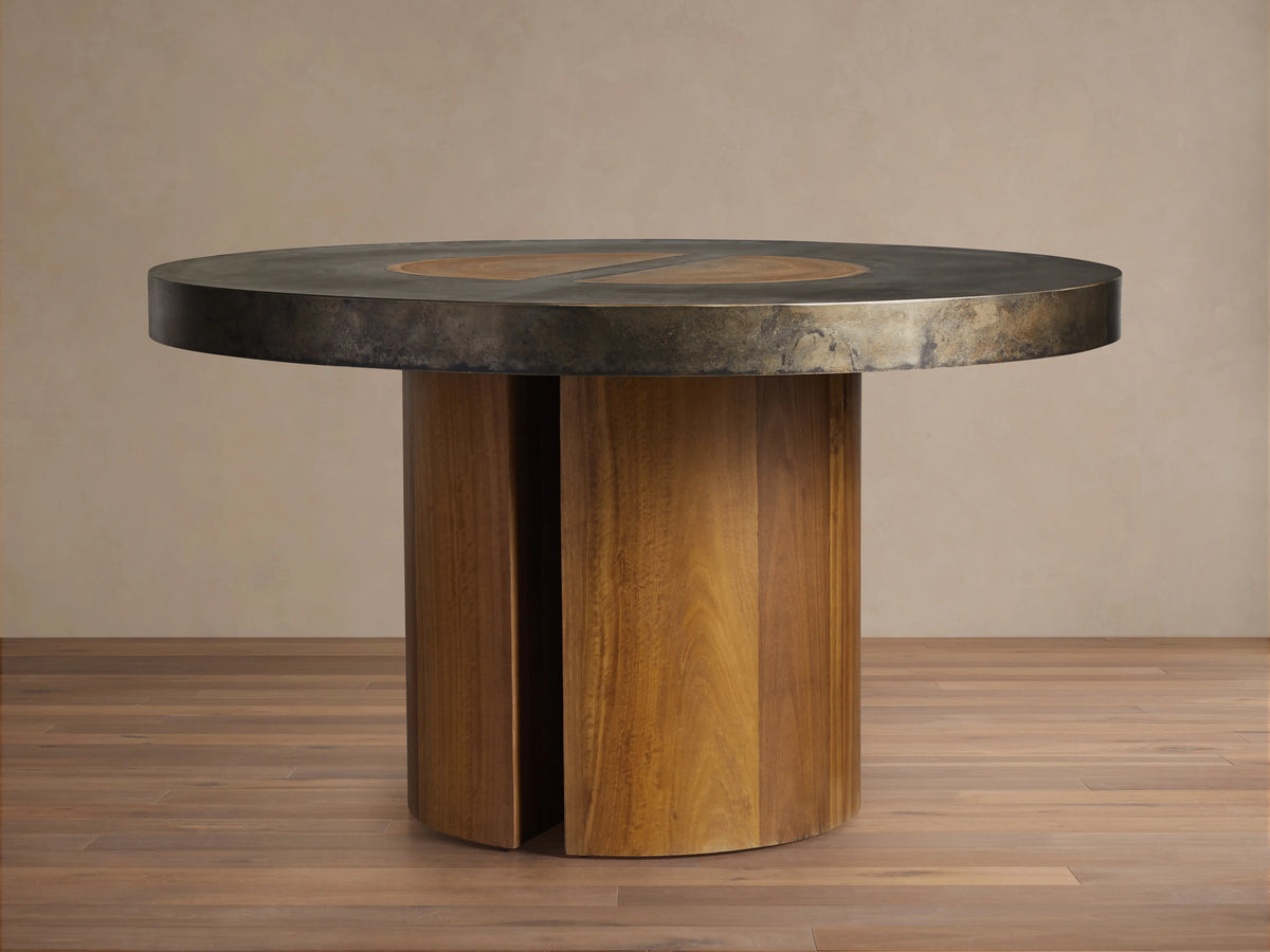 Acacius Round Dining Table with a dark round top featuring oxidized steel and yukas wood, set on a sturdy wooden pedestal base.