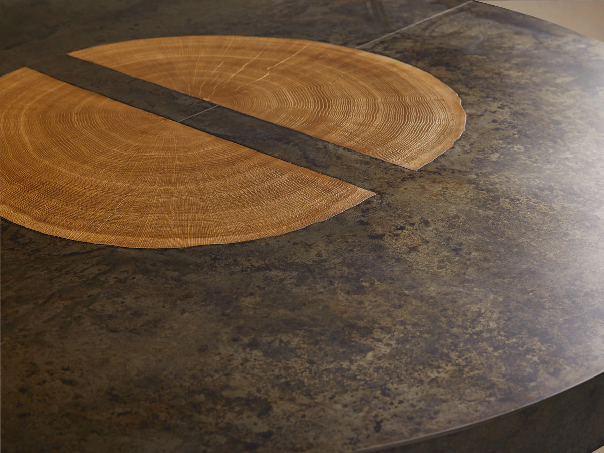 Round dining table with a dark, oxidized steel top, wooden veneer accents, and a base of solid wood panels on a wooden floor.