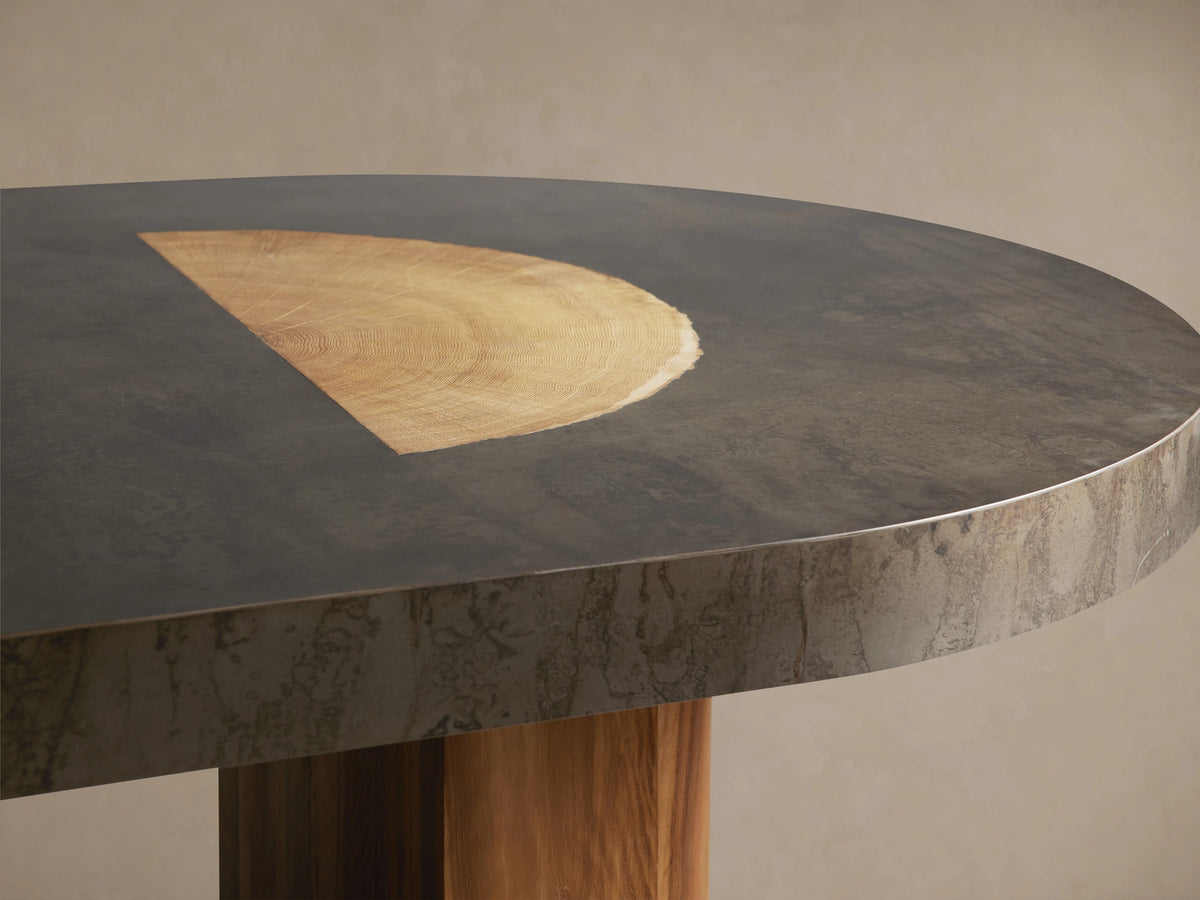 Oval dining table with a dark, weathered top made of oxidized steel and wood. Supported by two thick, rectangular wooden legs.