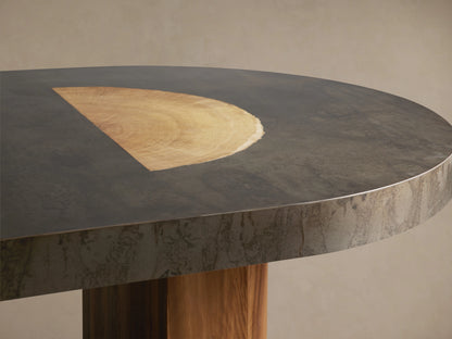 Oval dining table with a dark, weathered top made of oxidized steel and wood. Supported by two thick, rectangular wooden legs.