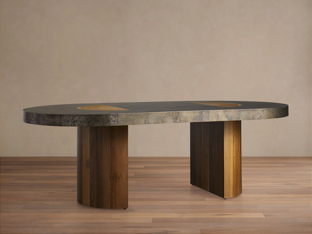 Acacius Dining Table with an oval top featuring oxidized steel and reclaimed yukas veneer. Two sturdy wooden legs support it.