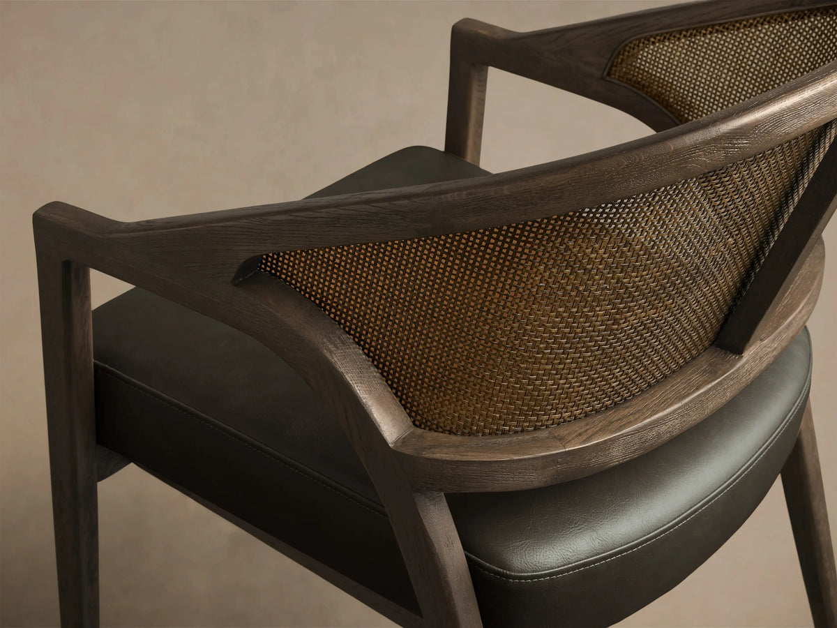 Side profile of the Black Drifted Aimee Faux Leather Dining Arm Chair, showing its dark oak frame, woven rattan cane back, and dark faux leather seat.