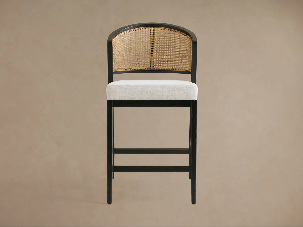 Aimee Armless Counter Stool with a solid oak frame, rattan cane back, and upholstered seat in light fabric on wooden floor.