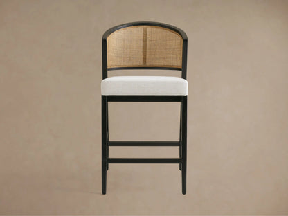 Aimee Armless Counter Stool with a solid oak frame, rattan cane back, and upholstered seat in light fabric on wooden floor.