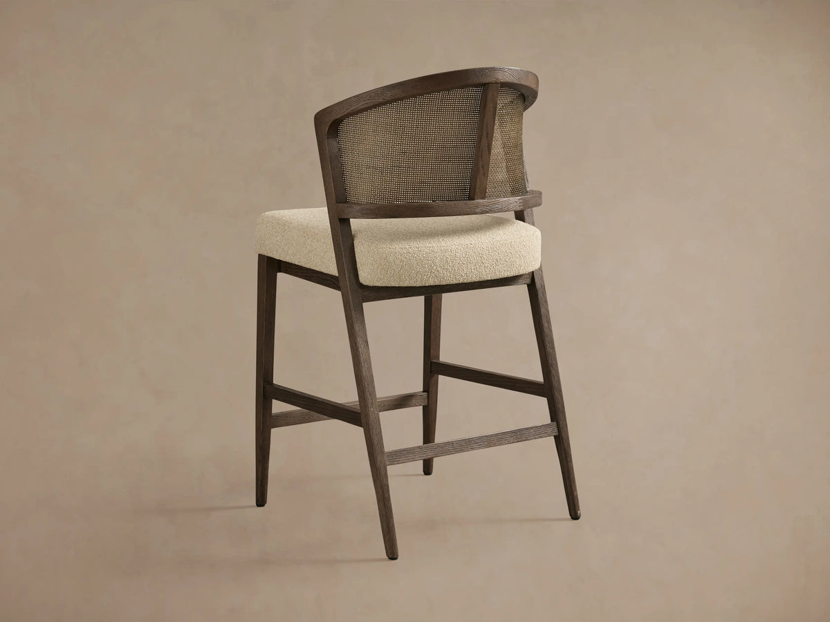 Aimee Armless Counter Stool with a solid oak frame and rattan cane back, upholstered in light fabric, on a neutral background.