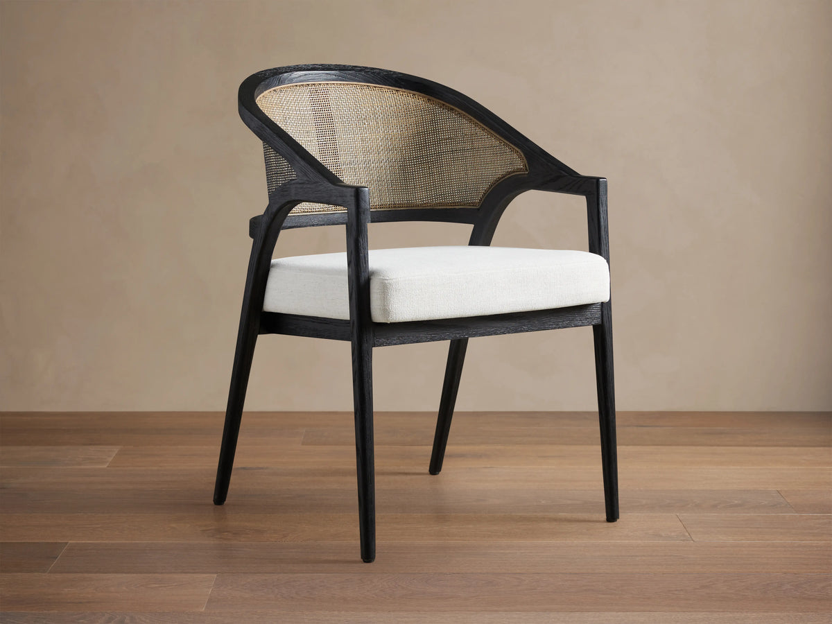Aimee Dining Arm Chair with black oak frame, rattan cane back, and a cream upholstered cushion. Set on wooden floor.