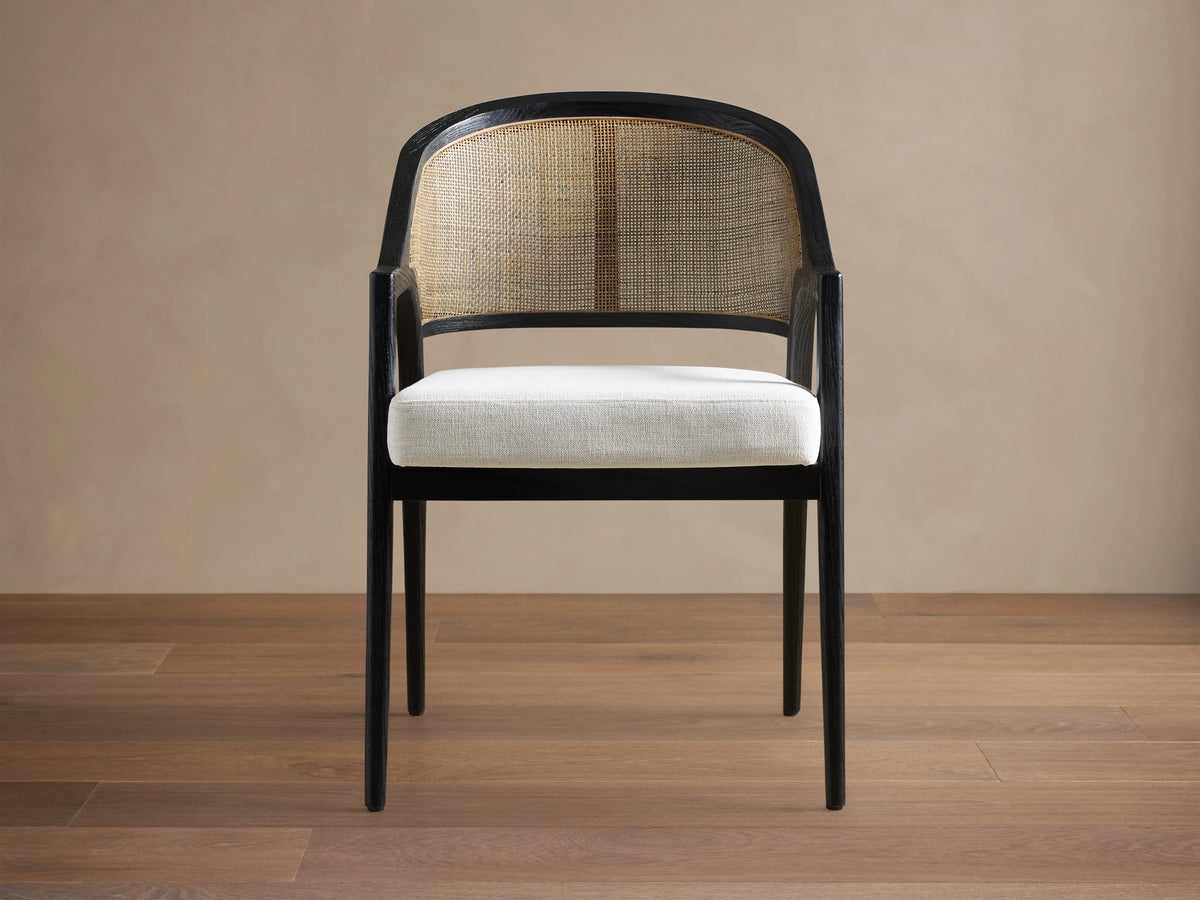 Front view of the Aimee Dining Arm Chair, featuring a dark brown solid oak frame, natural rattan cane back, and light beige upholstered cushion.