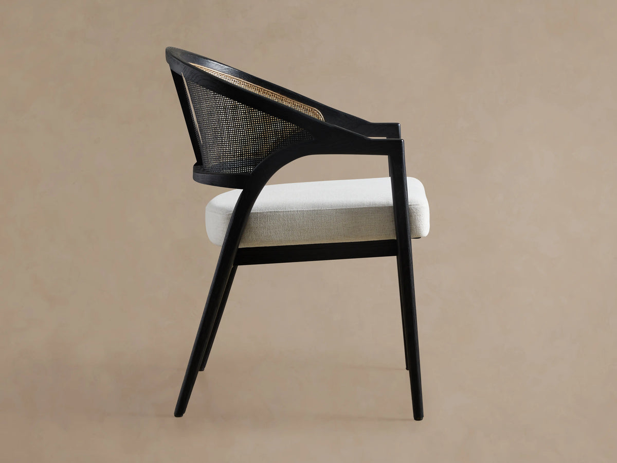 Aimee Dining Arm Chair with black drifted solid oak frame, rattan cane back, and white upholstered cushion seat, angled view.