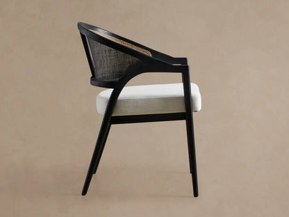 Aimee Dining Arm Chair with black drifted solid oak frame, rattan cane back, and white upholstered cushion seat, angled view.