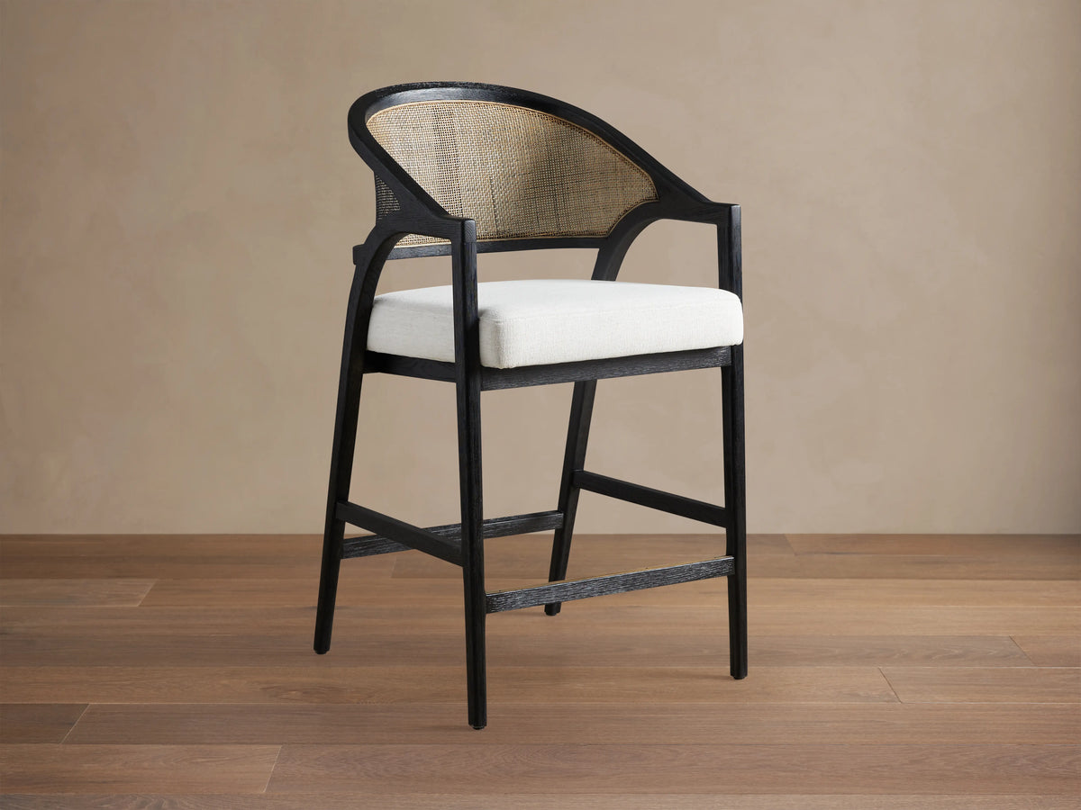 Aimee Stool with solid oak frame in Black Drifted finish, rattan cane back, and upholstered cream seat, on wooden floor.