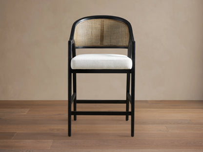 Aimee Stool with solid oak frame in Black Drifted finish, rattan cane back, and upholstered cream seat, on wooden floor.