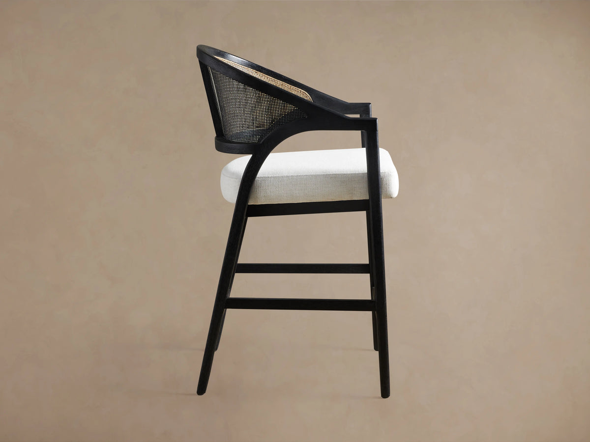 A black Aimee Stool with a rattan cane back and a white upholstered seat, showing its side profile on a beige background.