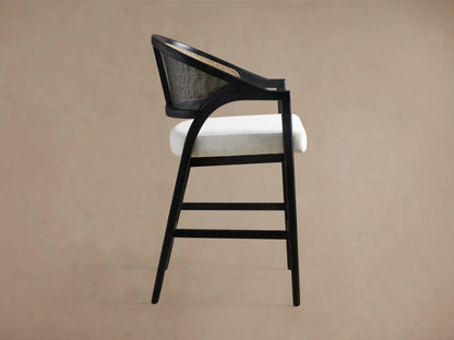 A black Aimee Stool with a rattan cane back and a white upholstered seat, showing its side profile on a beige background.