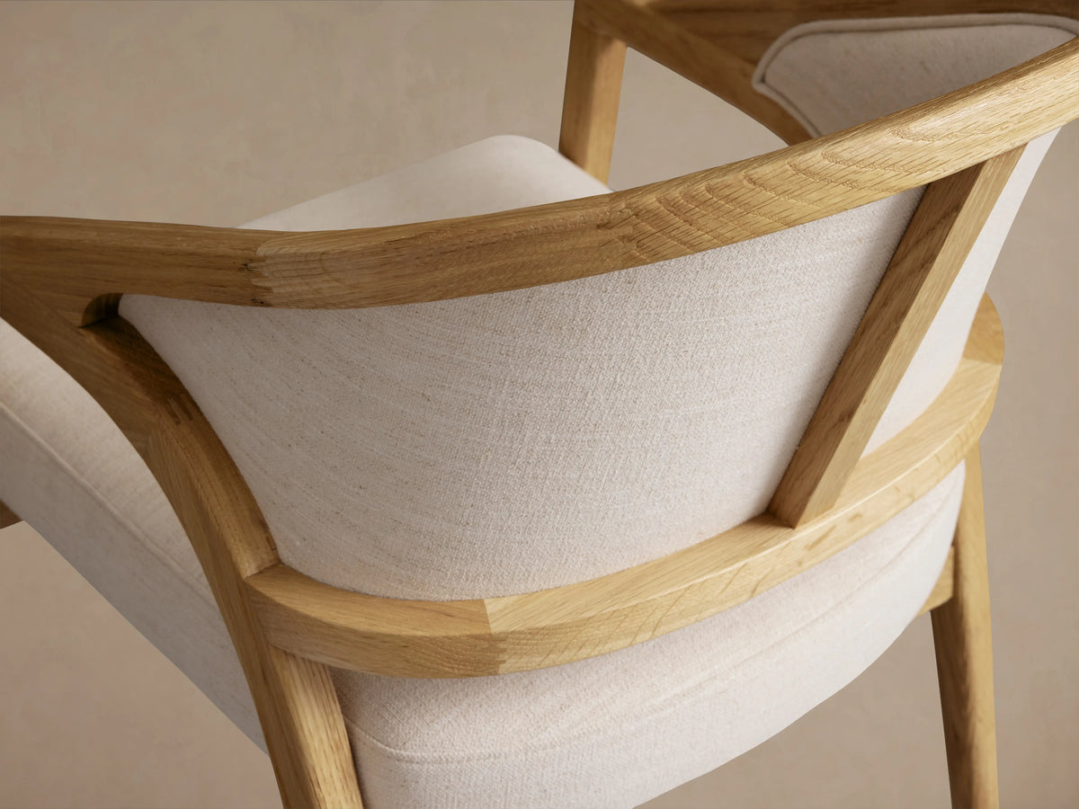 Aimee Upholstered Back Dining Arm Chair with solid oak frame, curved back, and cushioned seat in light fabric on wood floor.