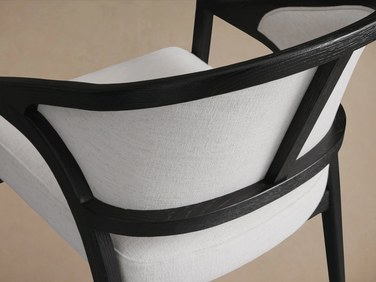 Aimee Upholstered Back Dining Arm Chair with a curved black oak frame and light beige fabric on seat and backrest, on wooden floor.