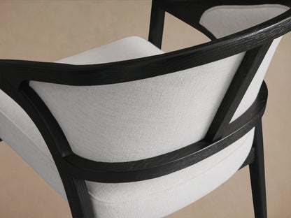 Aimee Upholstered Back Dining Arm Chair with a curved black oak frame and light beige fabric on seat and backrest, on wooden floor.