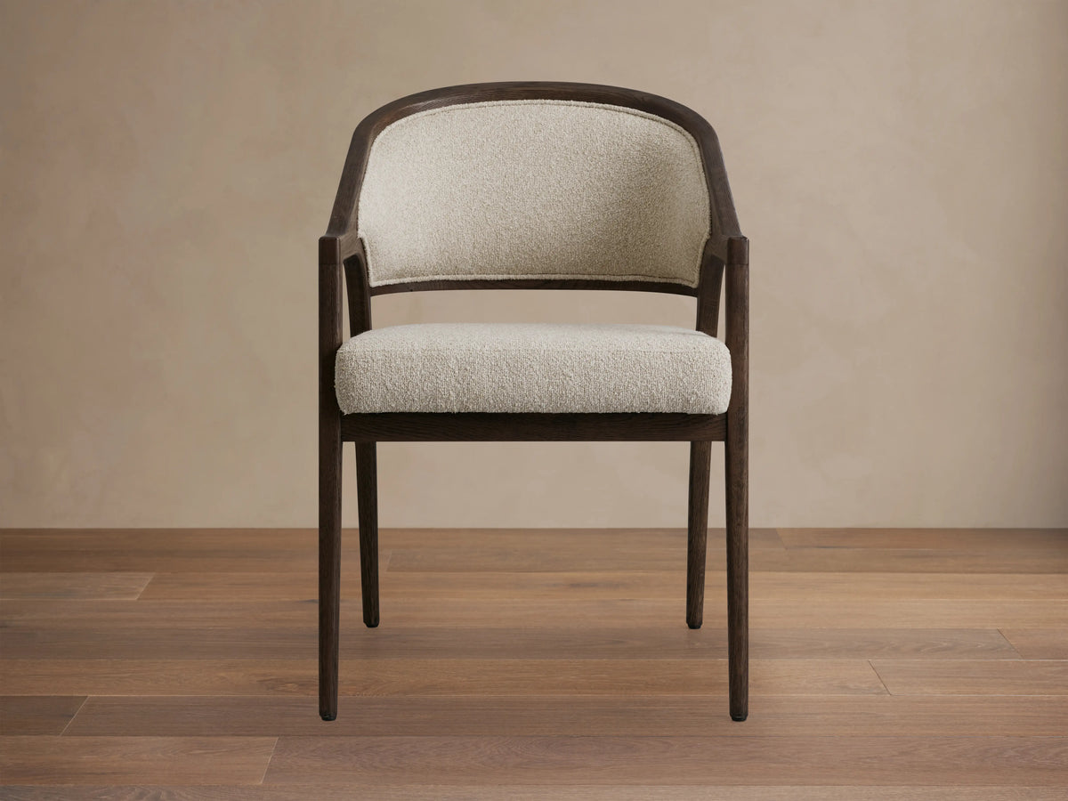 Side view of a dark brown Aimee Upholstered Back Dining Arm Chair with curved back, at a rustic wooden dining table.