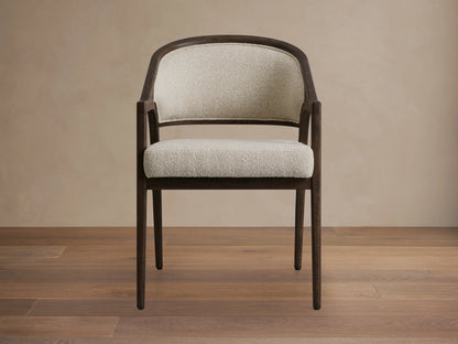 Side view of a dark brown Aimee Upholstered Back Dining Arm Chair with curved back, at a rustic wooden dining table.