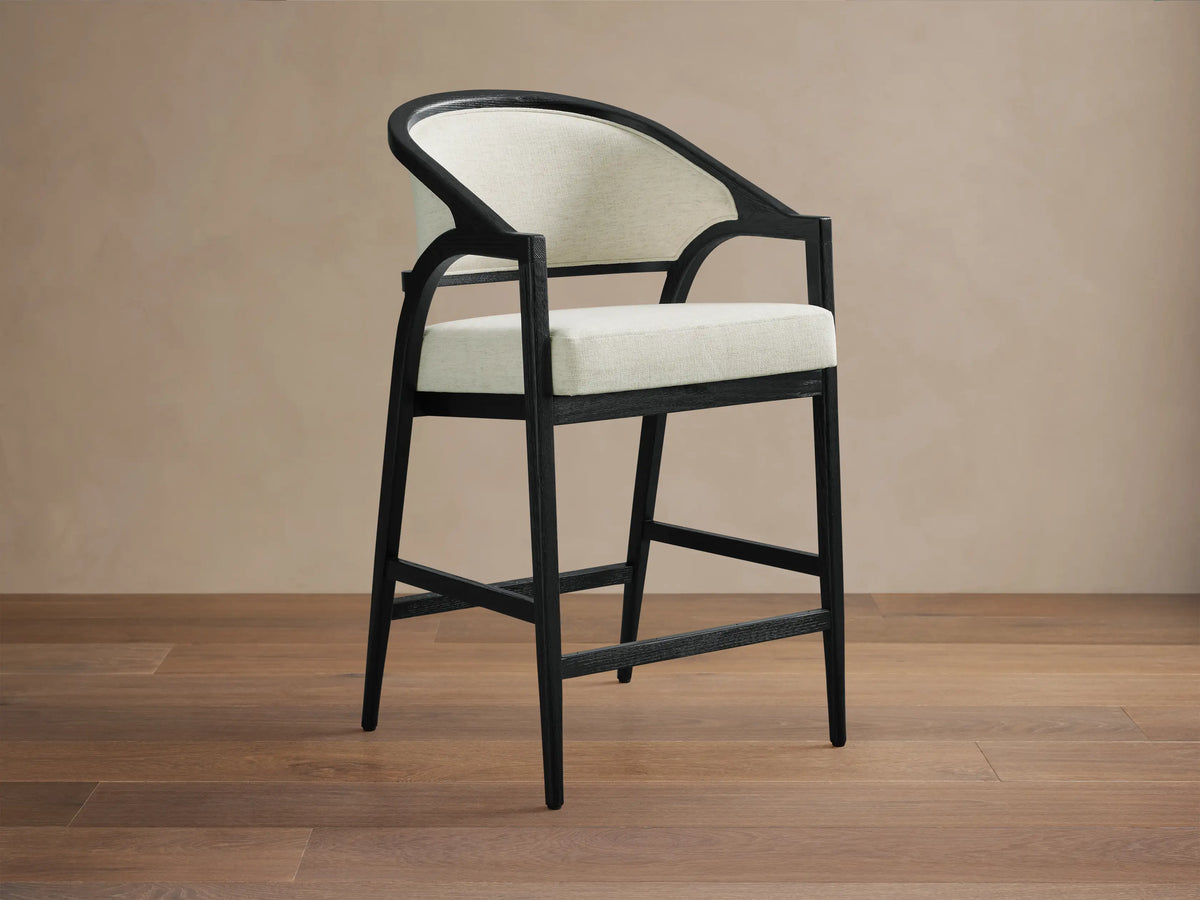 Aimee Upholstered Back Counter Stool with solid oak frame, black lacquer finish, cream upholstered back and seat, on wooden floor.
