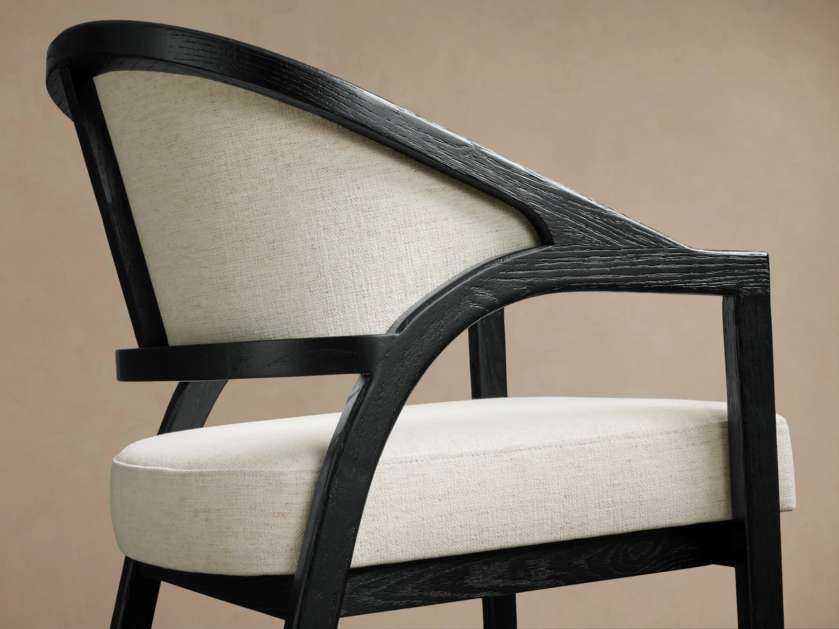 Aimee Upholstered Back Counter Stool with a black oak frame, beige fabric seat and backrest on a wooden floor background.