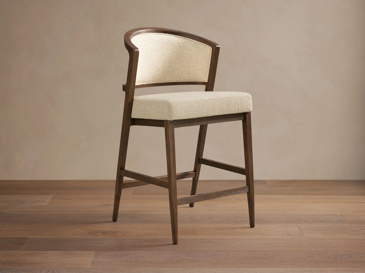 Aimee Upholstered Back Armless Counter Stool with a cream upholstered seat and back, solid oak frame, on a wooden floor.