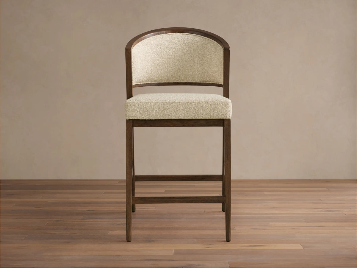 Aimee Upholstered Back Armless Counter Stool with a cream upholstered seat and back, solid oak frame, on a wooden floor.