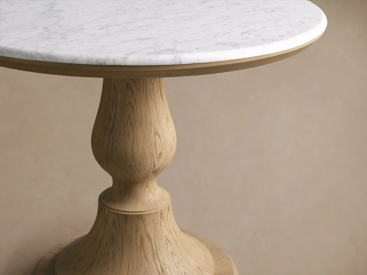 Amelia Bistro Table: round white marble top with gray veining, carved light oak pedestal base on a wooden floor.