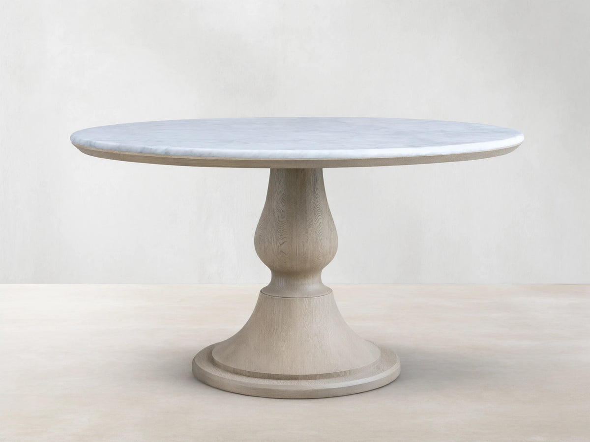 Amelia Dining Table: Round white marble top with gray veins, light oak pedestal base with visible wood grain, on wood floor.