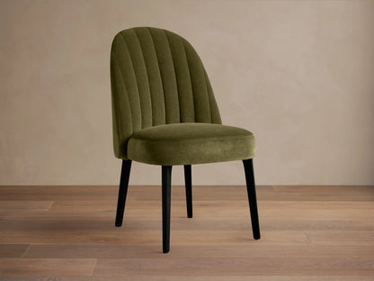 Arco Channel Dining Chair, olive green velvet with curved, channel-tufted back and black legs, angled on wood floor.