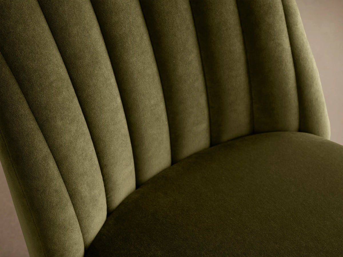 Frontal view: Arco Channel Dining Chair in olive green velvet with vertical channel tufting on the backrest, black legs, on a wooden floor.