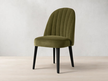 Arco Channel Dining Chair, olive green velvet with curved, channel-tufted back and black legs, angled on wood floor.