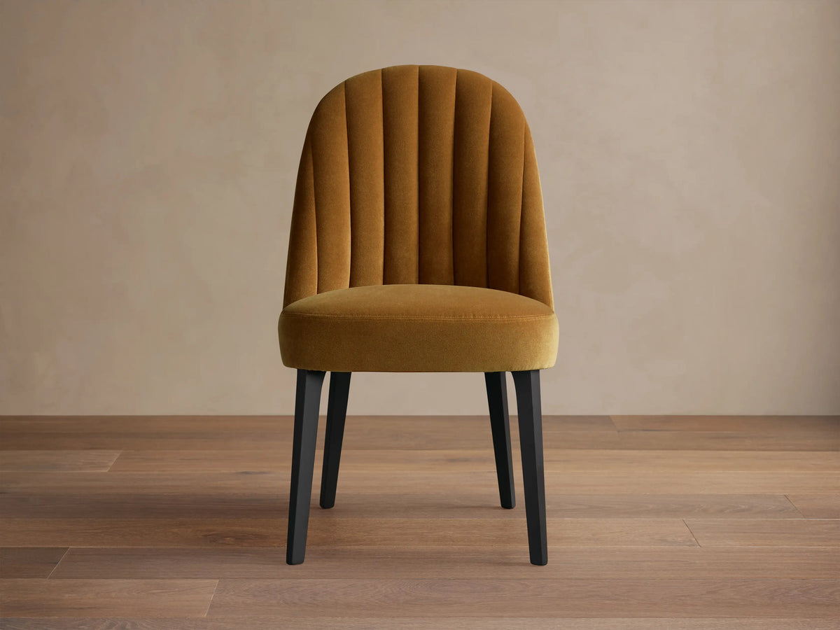 Arco Channel Dining Chair, mustard gold velvet with channel-tufted rounded back, padded seat, and sleek black legs on wood floor.