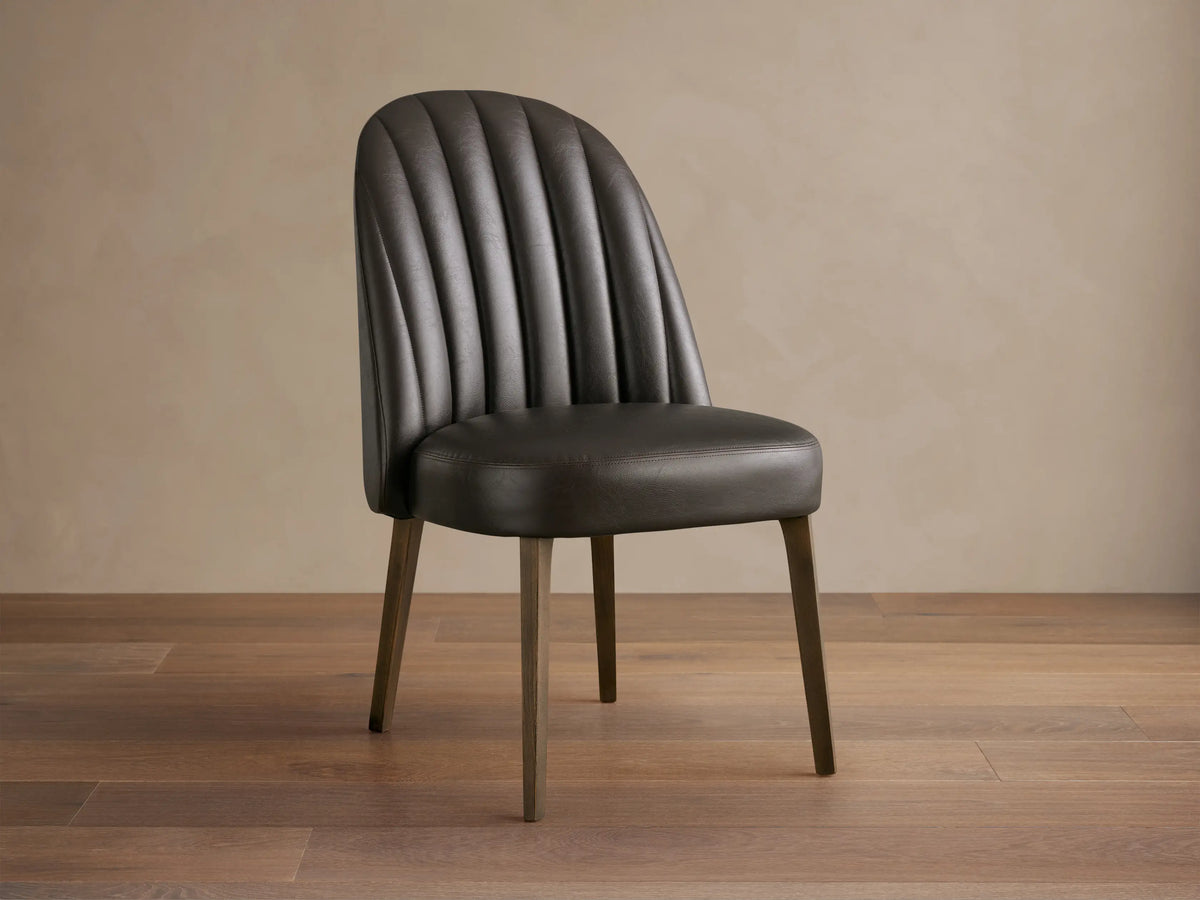Arco Dining Chair, dark brown faux leather with channel tufted curved backrest, padded seat, and tapered wood legs on a wood floor.