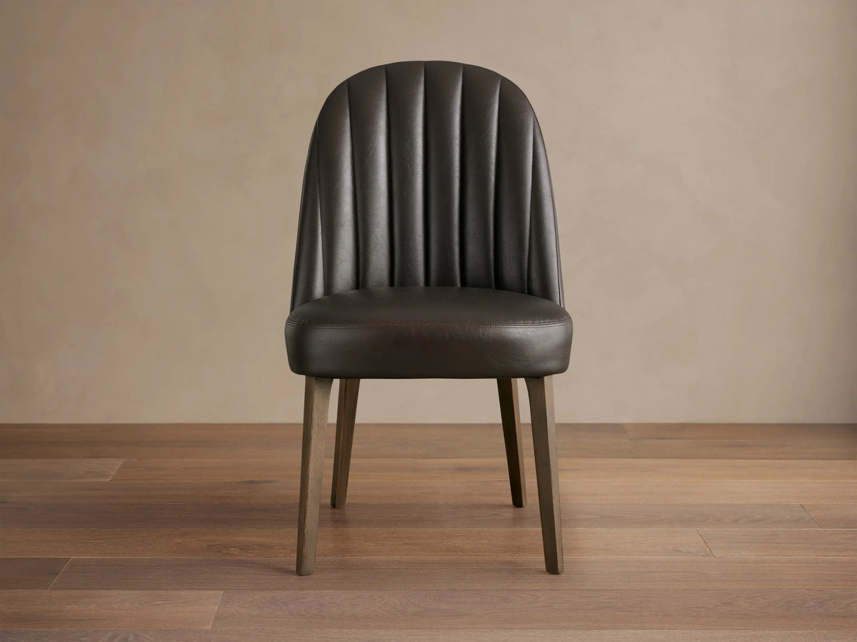 Arco Dining Chair, dark brown faux leather with channel tufted curved backrest, padded seat, and tapered wood legs on a wood floor.