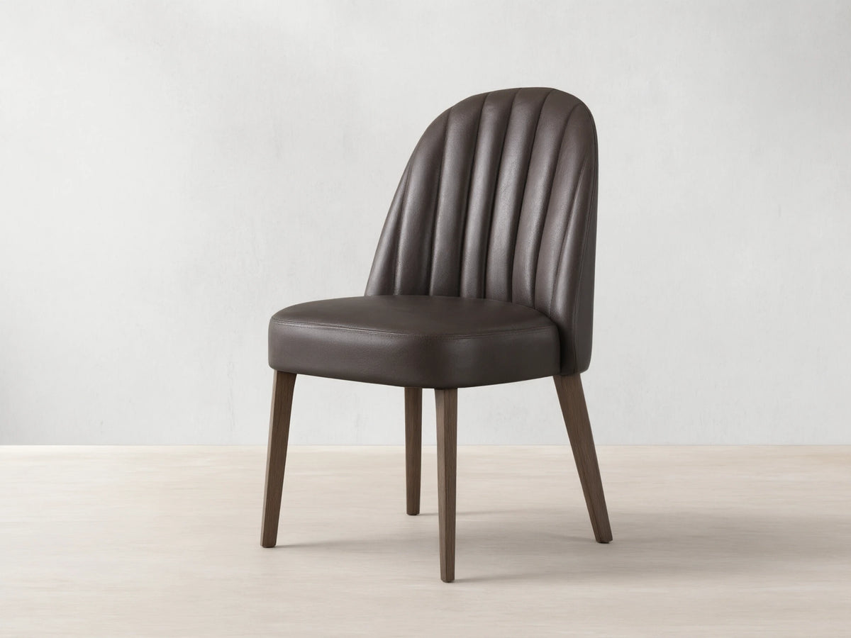Arco Dining Chair, dark brown faux leather with channel tufted curved backrest, padded seat, and tapered wood legs on a wood floor.
