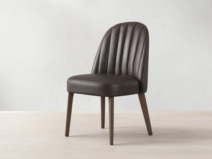 Arco Dining Chair, dark brown faux leather with channel tufted curved backrest, padded seat, and tapered wood legs on a wood floor.