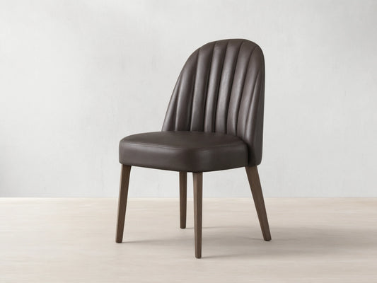 Arco Dining Chair, dark brown faux leather with channel tufted curved backrest, padded seat, and tapered wood legs on a wood floor.