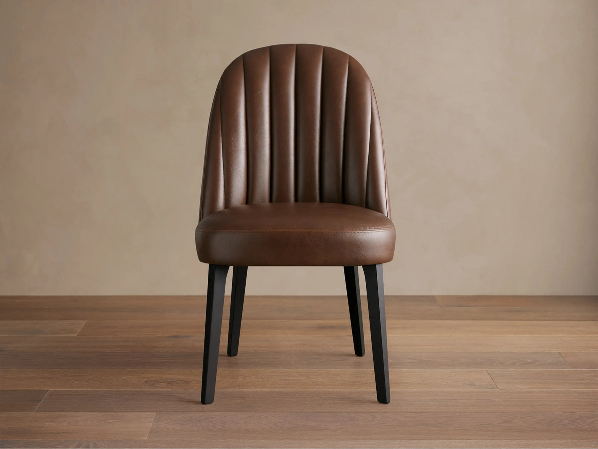 Arco Faux Leather Dining Chair: Brown, channel-tufted curved back, dark legs. Centered on a wooden floor.