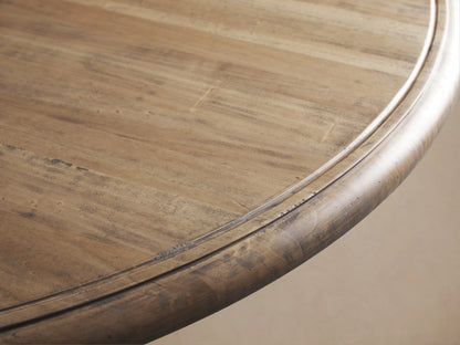 Round Asolo Dining Table with a thick pedestal base, antique finish, and hand-carved detailing on wooden flooring in a light-toned room.