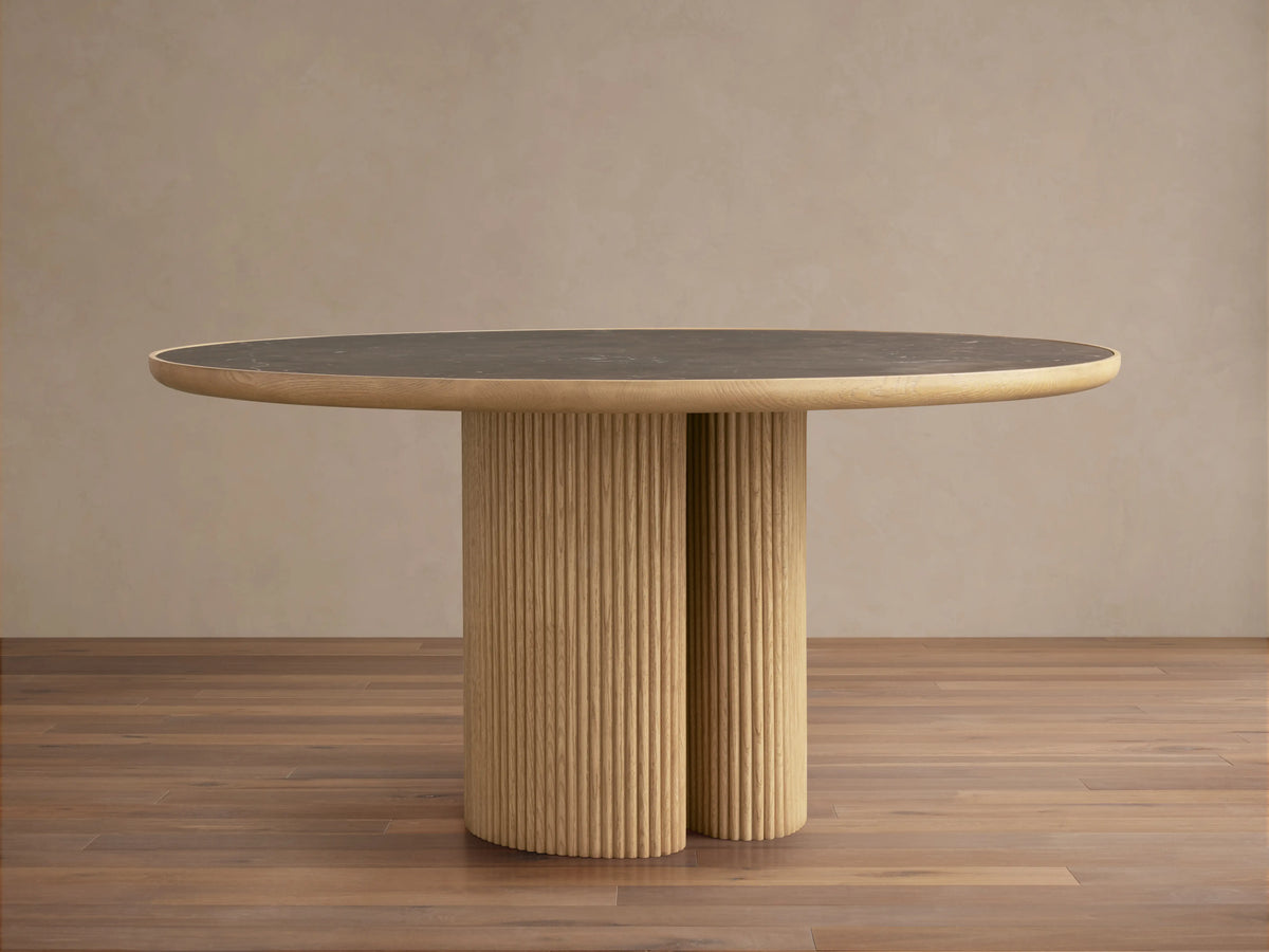 Astor Round Dining Table with a dark marble top and ribbed oak wood base, set on a wooden floor in a neutral room.