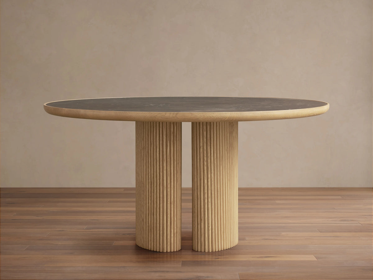 Astor Round Dining Table with a dark marble top and ribbed oak wood base, set on a wooden floor in a neutral room.